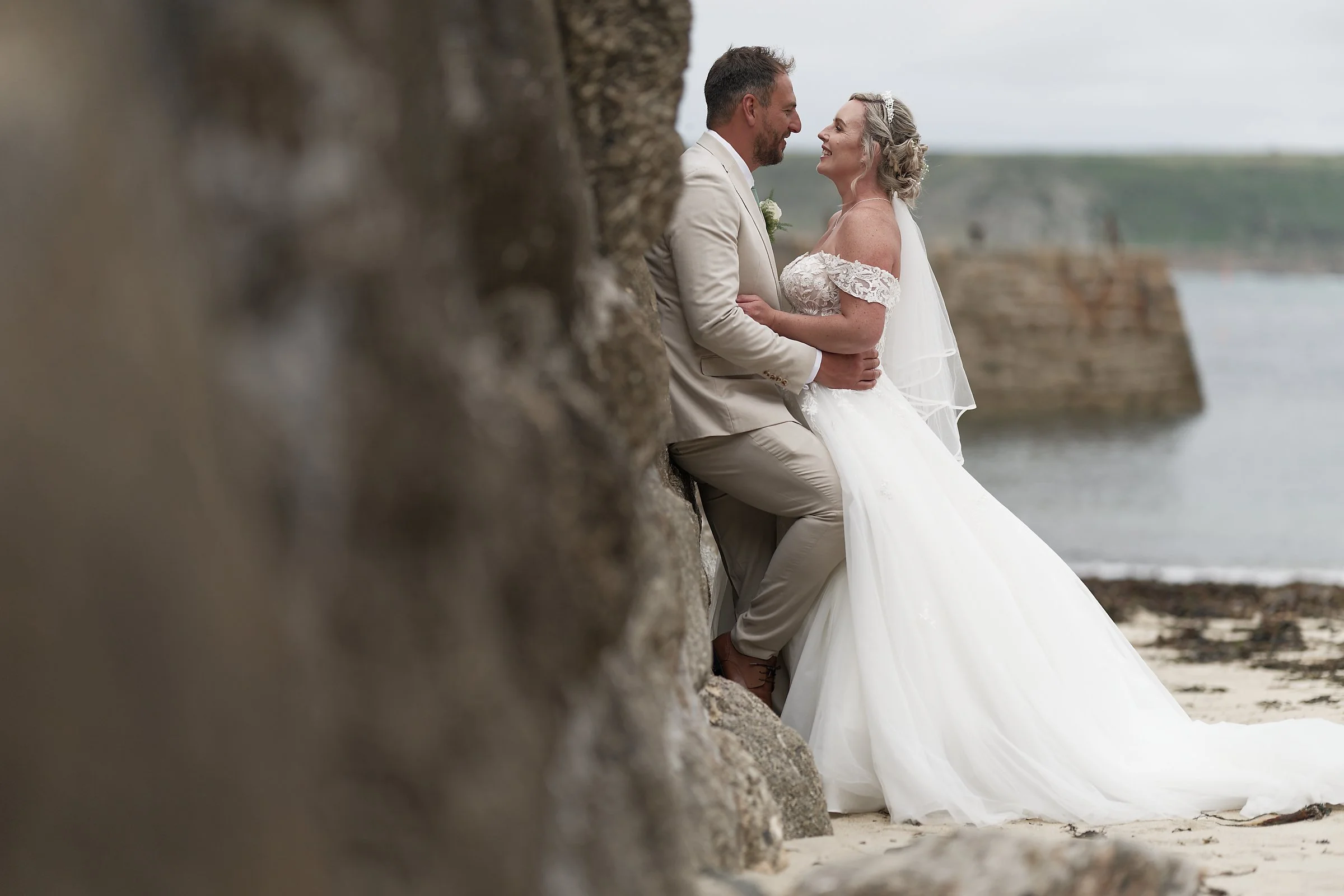 A couple dressed in wedding attire stands close together on a beach, smiling and gazing into each other's eyes, with wedding vows possibly being exchanged.