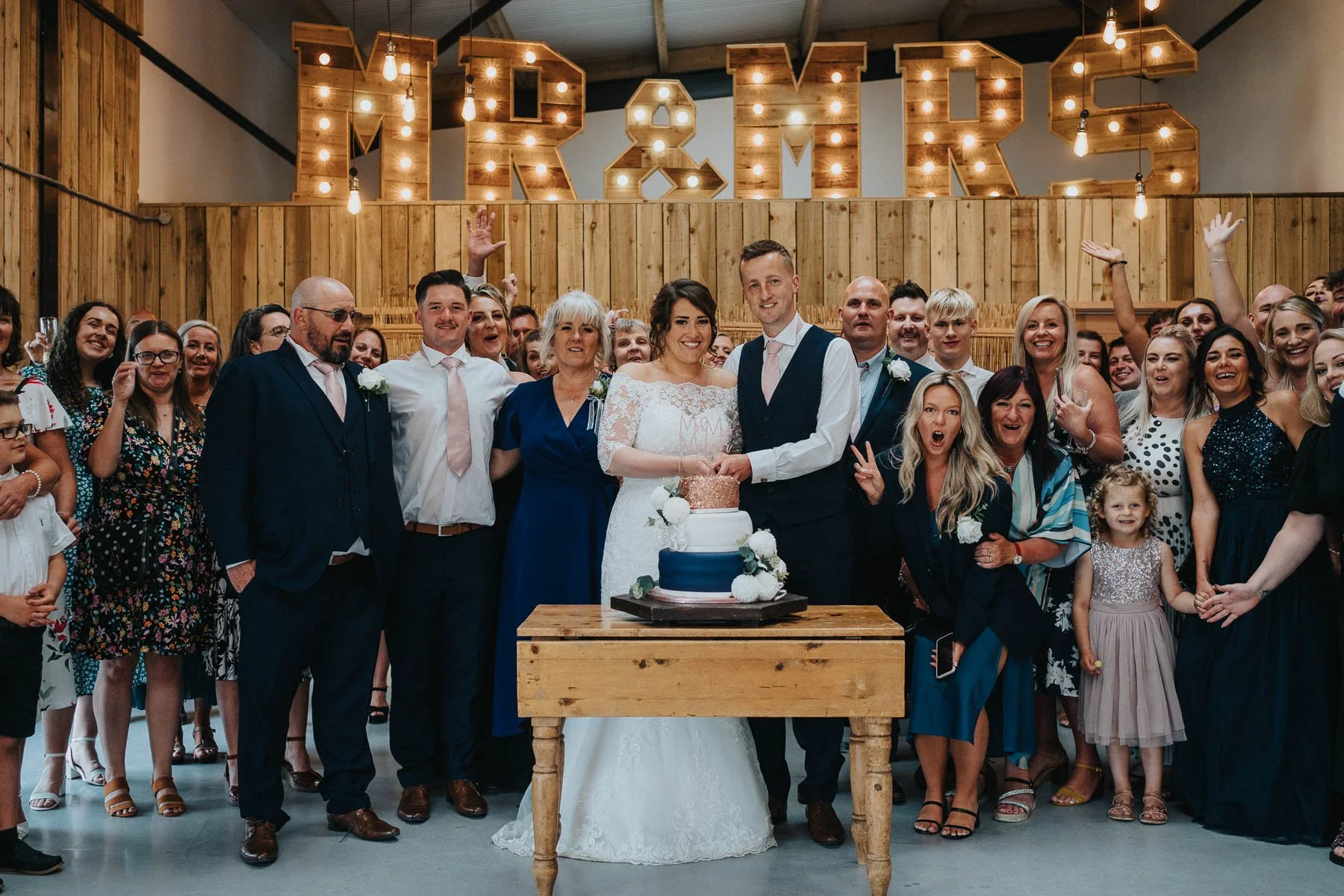 A large group of wedding guests celebrating around a bride and groom who are cutting a wedding cake, with a wooden backdrop and illuminated marquee letters spelling 'MRS' in the background.