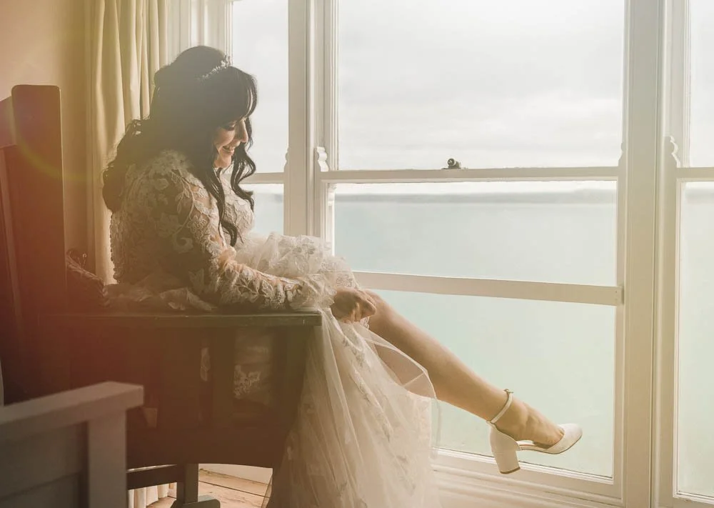 Bride sitting by a window, wearing a lace wedding dress and white high heels, smiling and looking down.