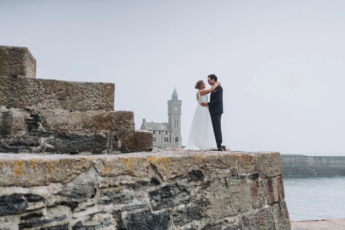A bride and groom embrace on a stone ledge by the water with a historic clock tower in the background.