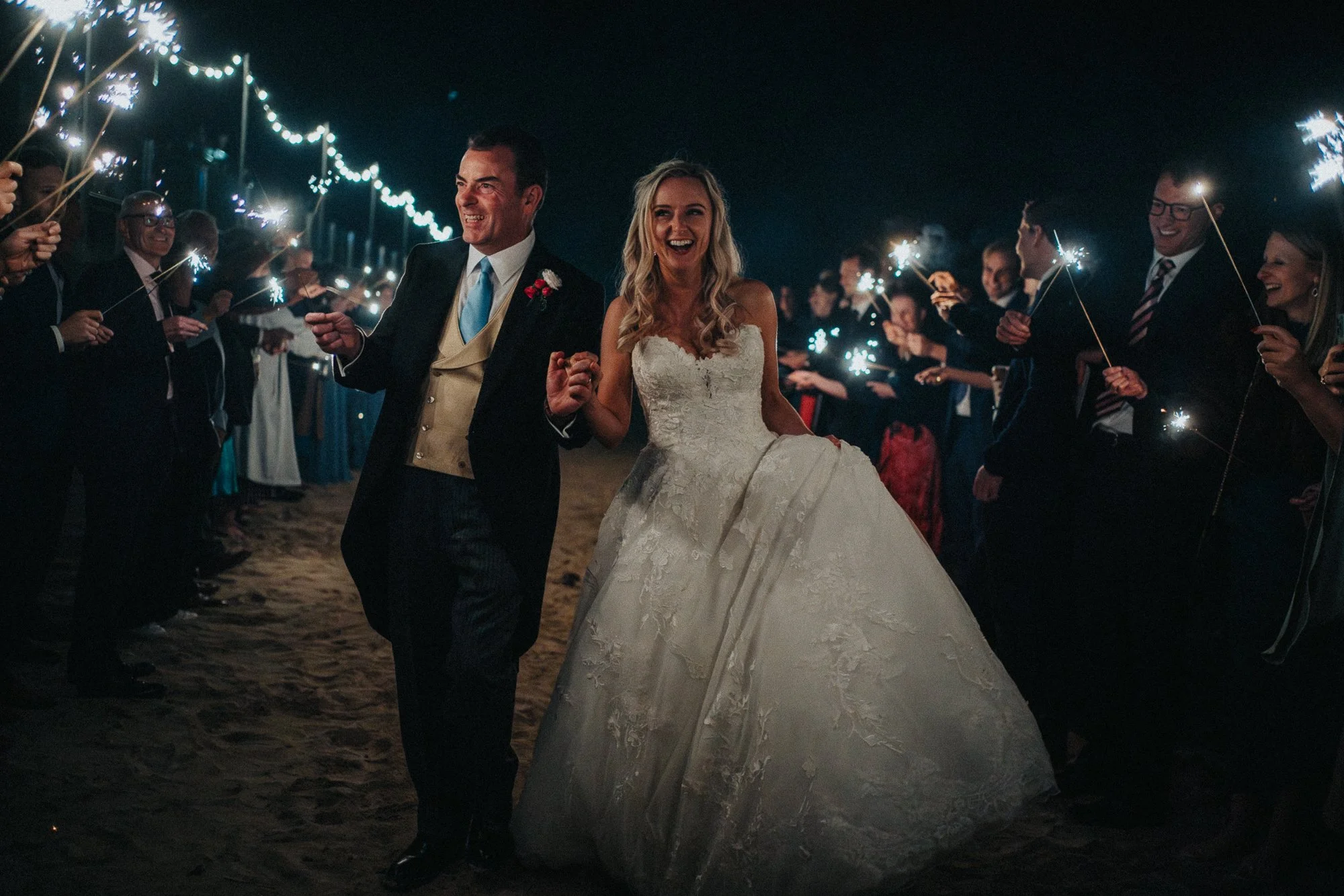 Bride and groom walking on the beach at night surrounded by guests holding sparklers.