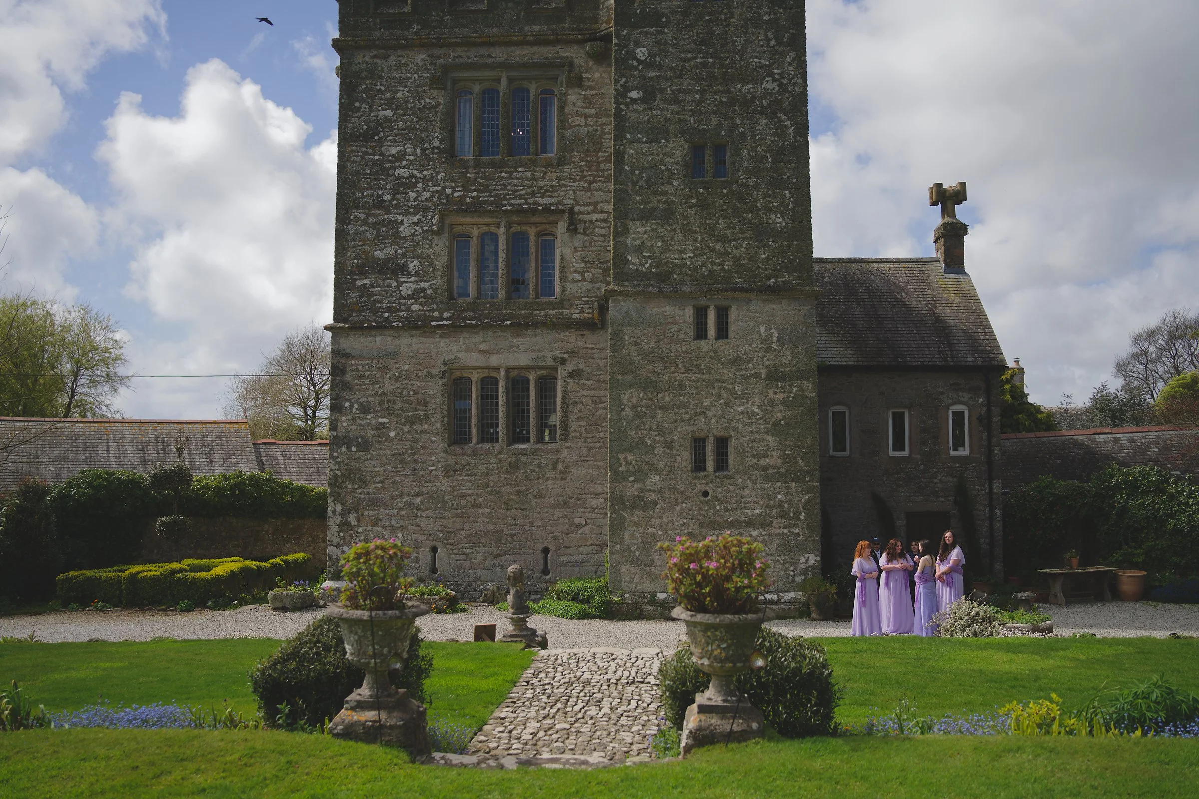 A group of women dressed in purple or lavender gowns standing in front of an old stone church with gothic windows. The church has a weathered stone facade and a cross on the roof. The foreground features a landscaped garden with potted plants, a cobb