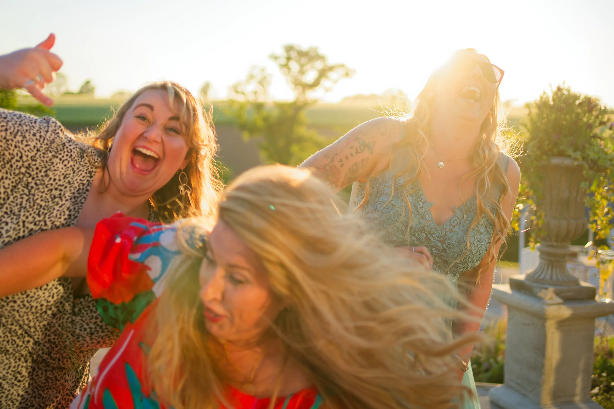 Two women and a child outdoors during sunset, smiling and playing together, with trees and a decorative stone garden sculpture in the background.