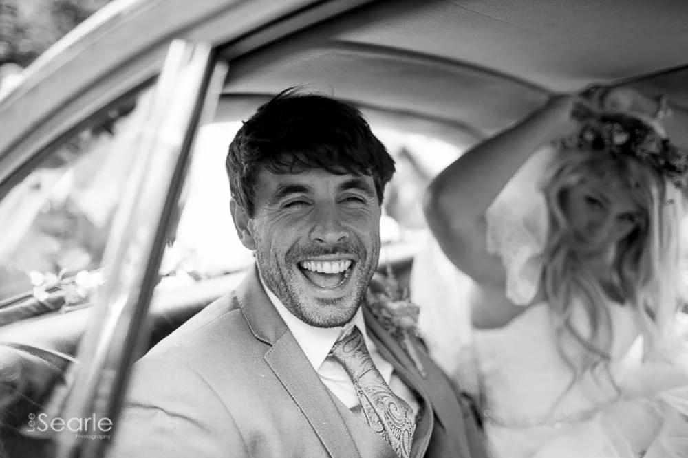 A happy man laughing inside a car with a woman who is adjusting a flower crown on her head.