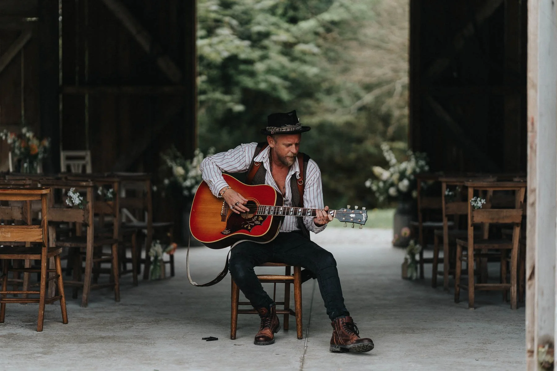 A man is sitting on a wooden chair, playing an acoustic guitar inside a rustic barn or shed decorated for a wedding or event.
