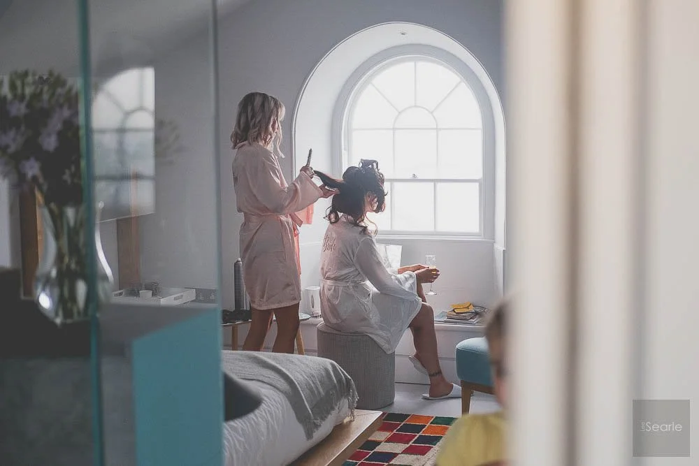 Two women in matching pink robes, one standing and styling the other’s hair, inside a bright, airy room with a large arched window in the background.