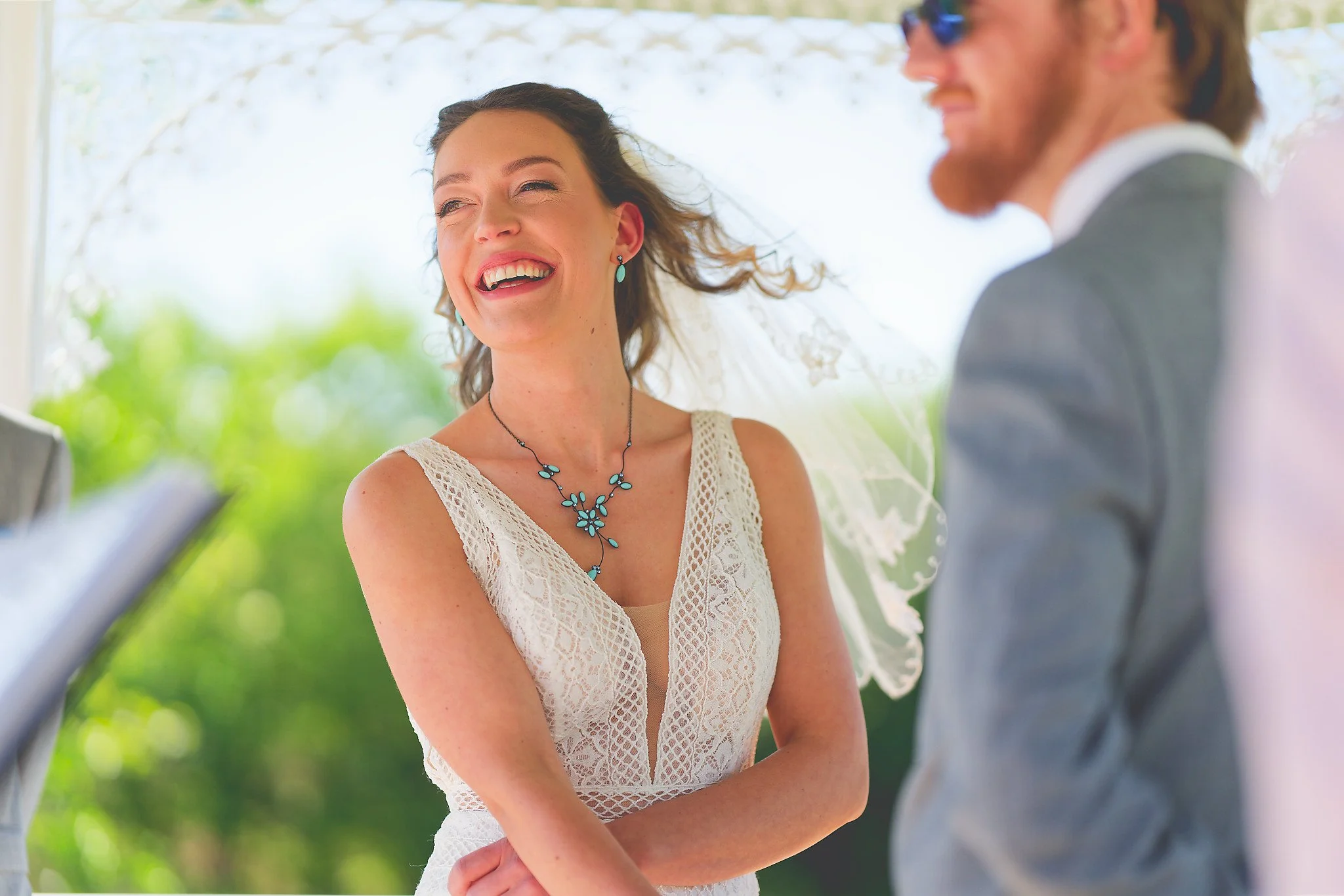 A woman in a white lace dress with a necklace and earrings, smiling and looking happy, during a wedding ceremony outdoors.