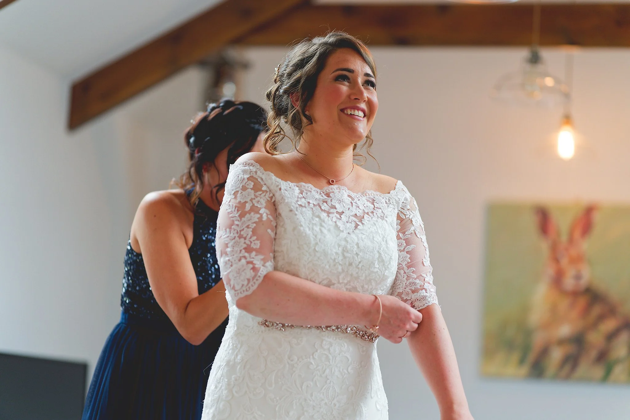 A bride in a white lace wedding dress smiling as a bridesmaid helps her prepare, with a painting of a rabbit on the wall in the background.