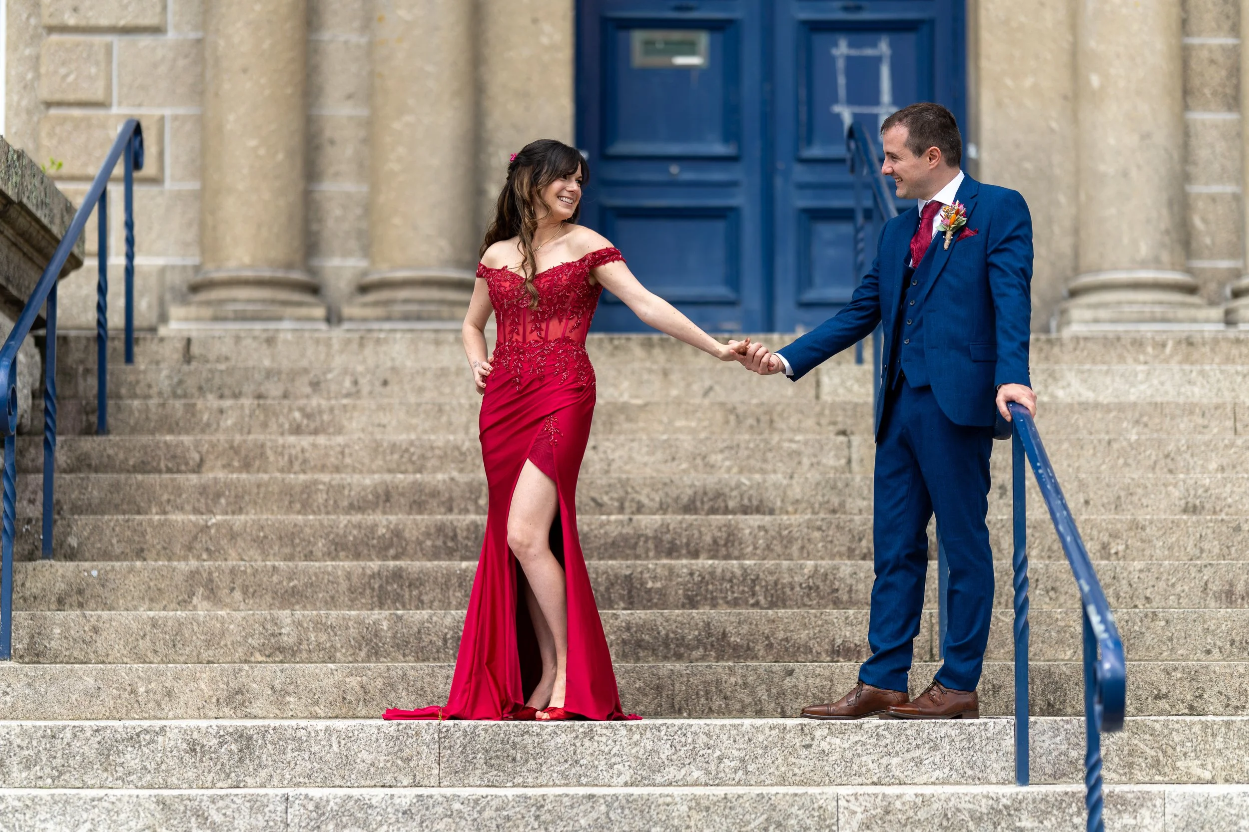 A woman in a red off-shoulder gown holding the hand of a man in a blue suit on a stone staircase in front of a church.
