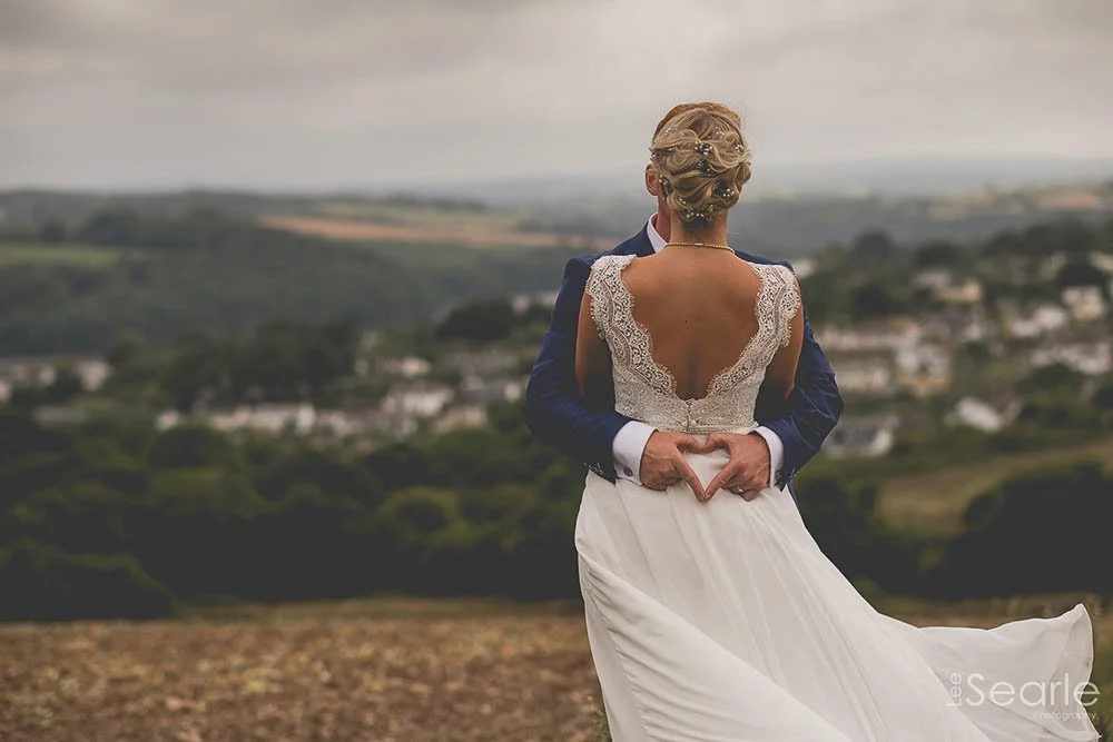 A bride and groom embracing outdoors, with the bride making a heart shape with her hands, overlooking a scenic landscape of fields and trees.