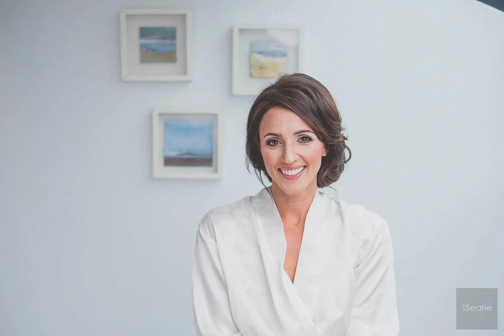 A woman with short brown hair smiling in a white blouse, standing in front of a white wall with three framed landscape paintings.