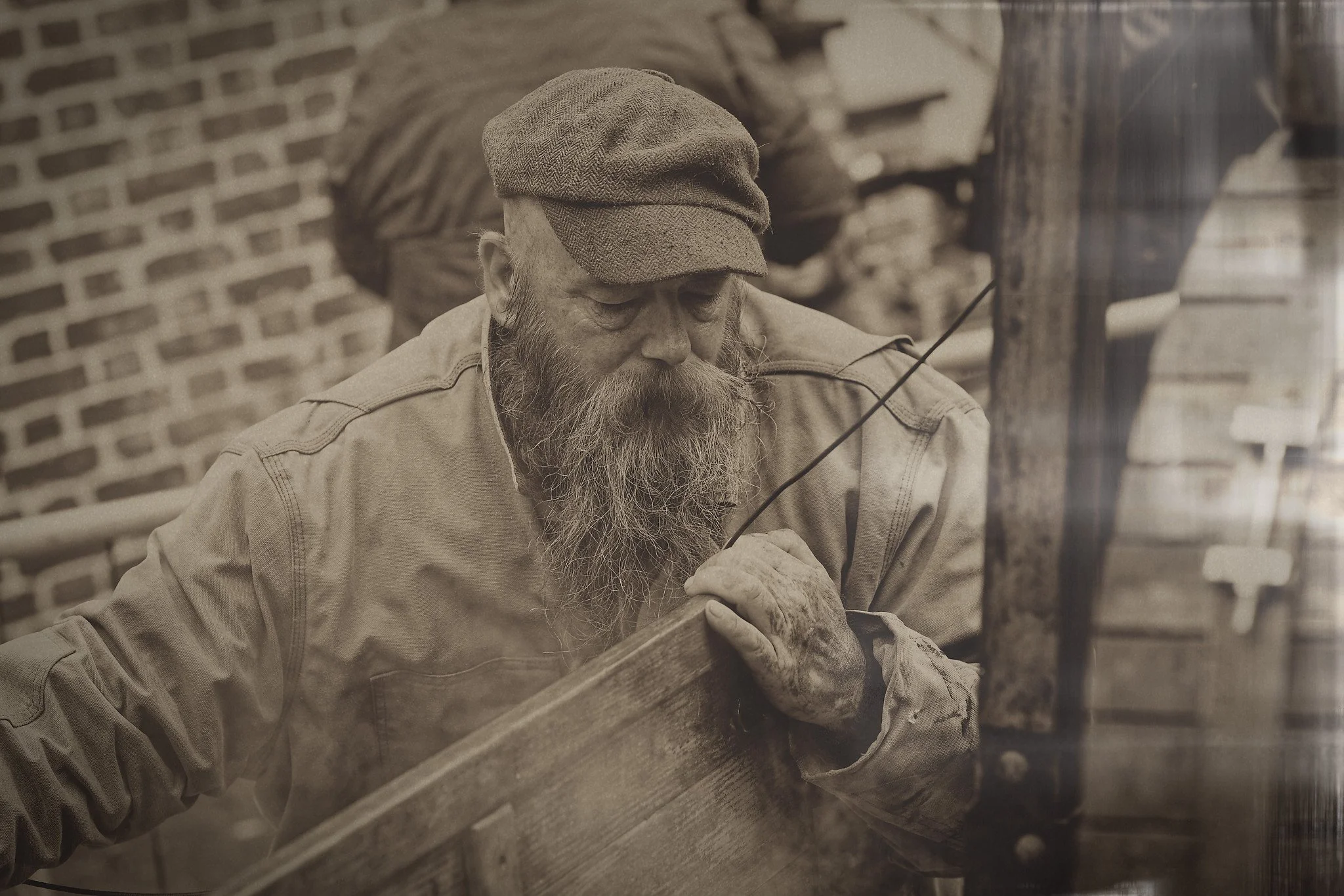 An elderly man with a long beard, wearing a cap and work shirt, working with wood outdoors near a brick wall.