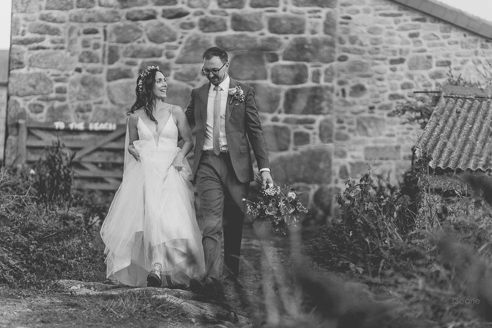 A bride and groom walking outdoors, smiling, with the bride holding the groom's hand. The bride wears a white wedding gown and floral headpiece; the groom is in a suit holding a bouquet. They walk along a stone path with a stone wall and plants in th