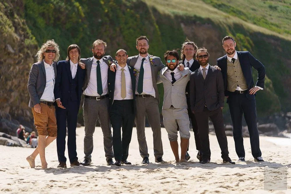 Group of nine people in suits is standing on the beach, smiling and embracing each other with a cliff in the background.