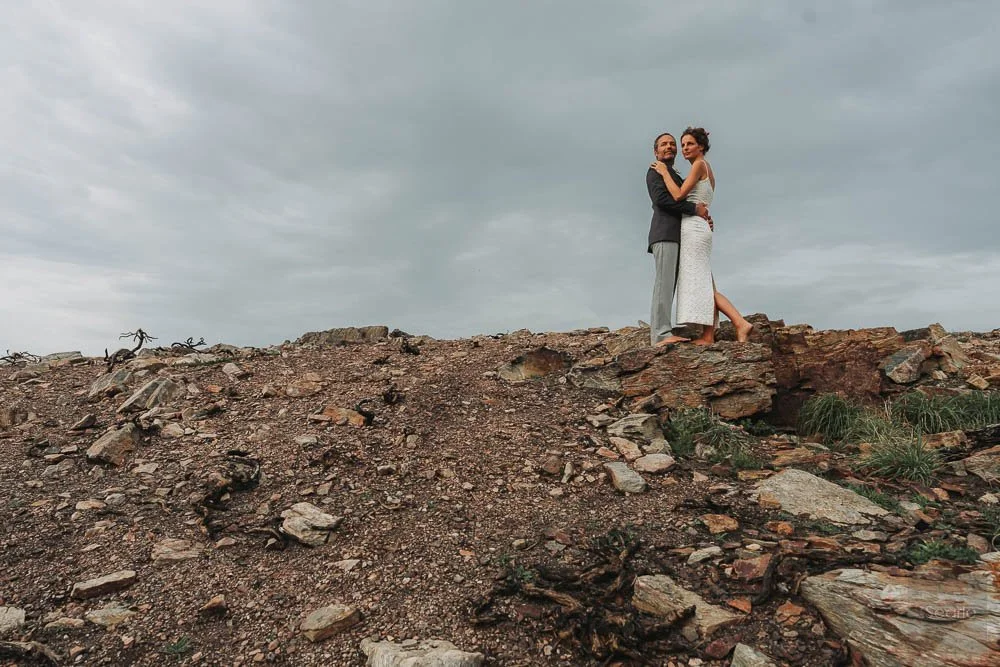 A couple dressed in formal attire standing on a rocky hilltop, embracing each other against a cloudy sky.