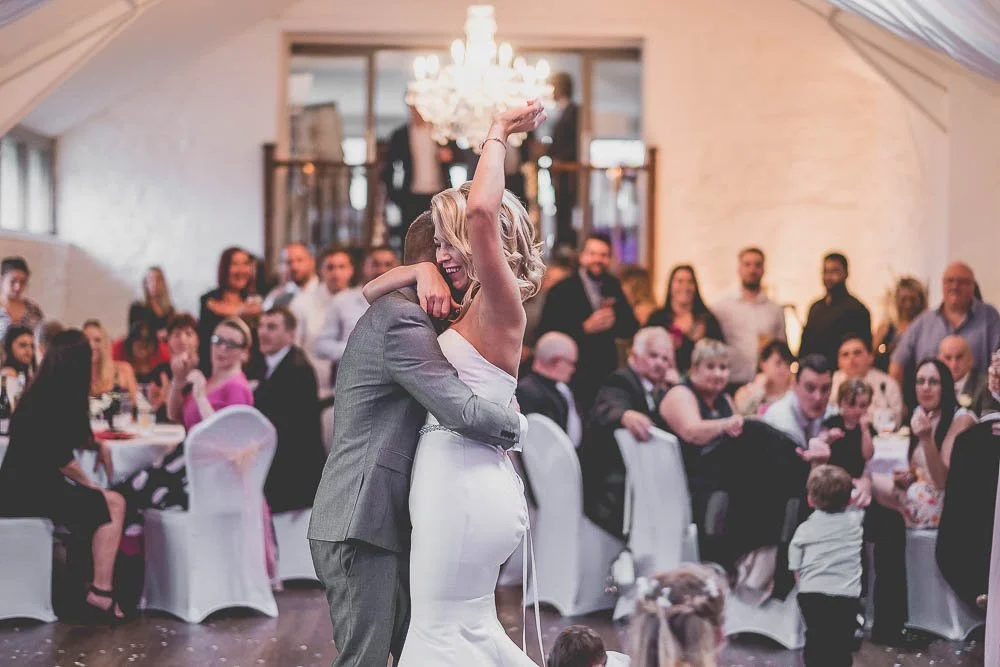 A bride and groom dance together in a wedding reception, surrounded by seated and standing guests in a decorated venue with a chandelier.