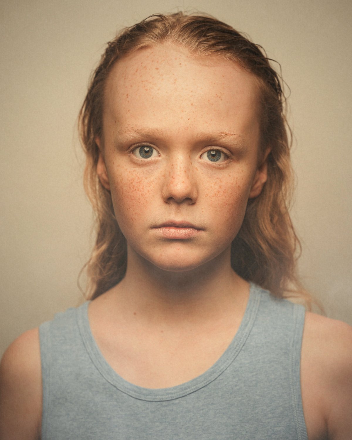 Close-up portrait of a young girl with red hair and blue eyes, wearing a gray sleeveless top, against a plain beige background.