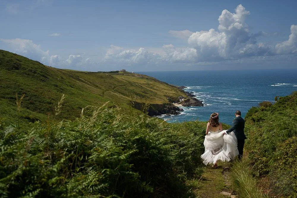 A bride and groom walking hand-in-hand on a lush green coastal trail towards the ocean, with cliffs and a partly cloudy sky in the background.