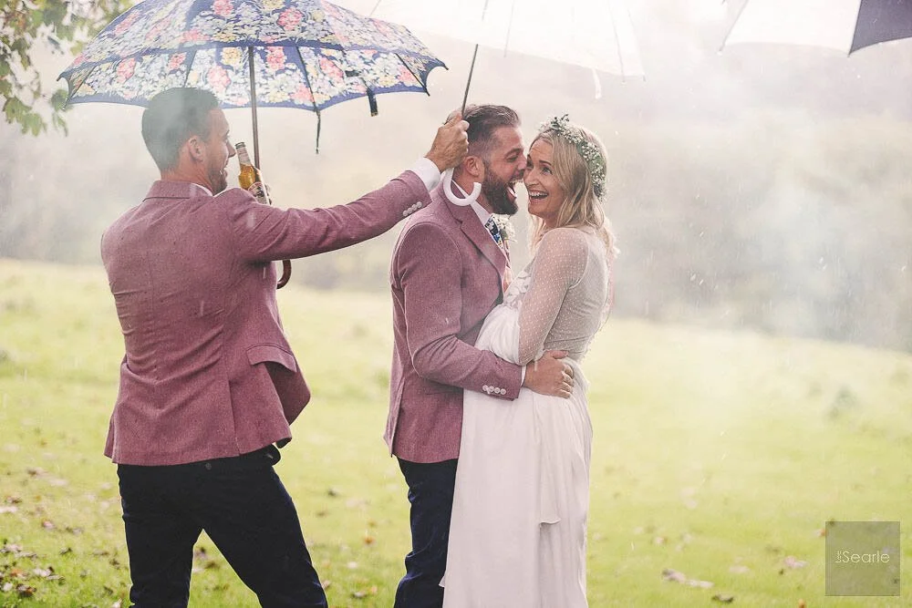 A joyful couple wedding photo with a man in a pink jacket holding a bride in a white dress and a floral headband, while a friend in a pink jacket holds an umbrella over them during a light rain.