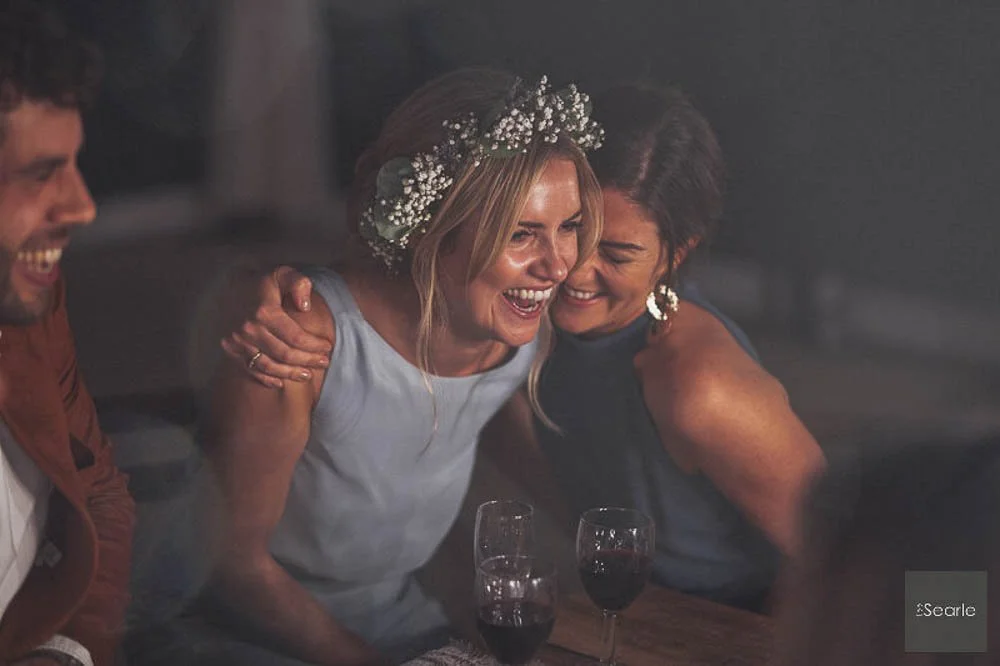 Three people at a table, smiling and laughing, with two glasses of red wine in front of them, celebrating indoors.