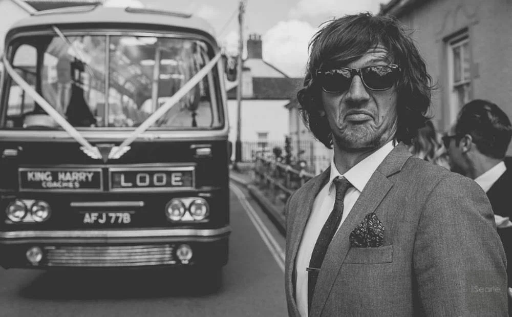 A man with wavy hair, wearing sunglasses, a suit, and a tie, making a funny face on a street with a bus labeled "King Harry Looe" parked behind him.