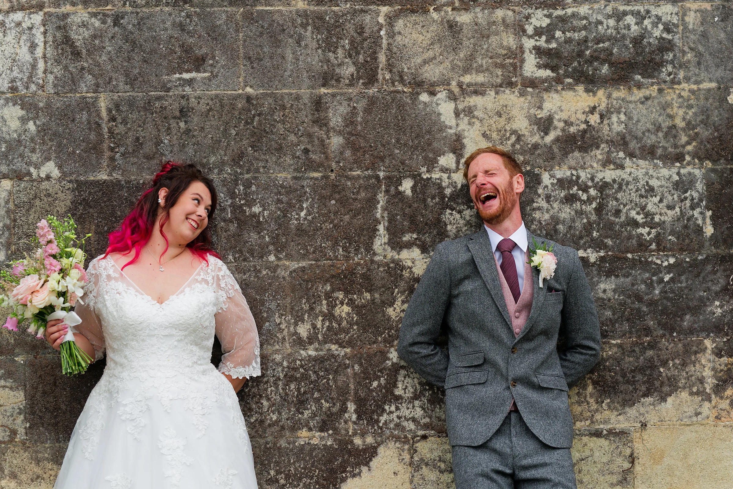 A bride and groom laughing and looking at each other while standing in front of a large stone wall. The bride is holding a bouquet of flowers and wearing a white lace wedding dress, and the groom is wearing a gray suit with a tie and a boutonniere.