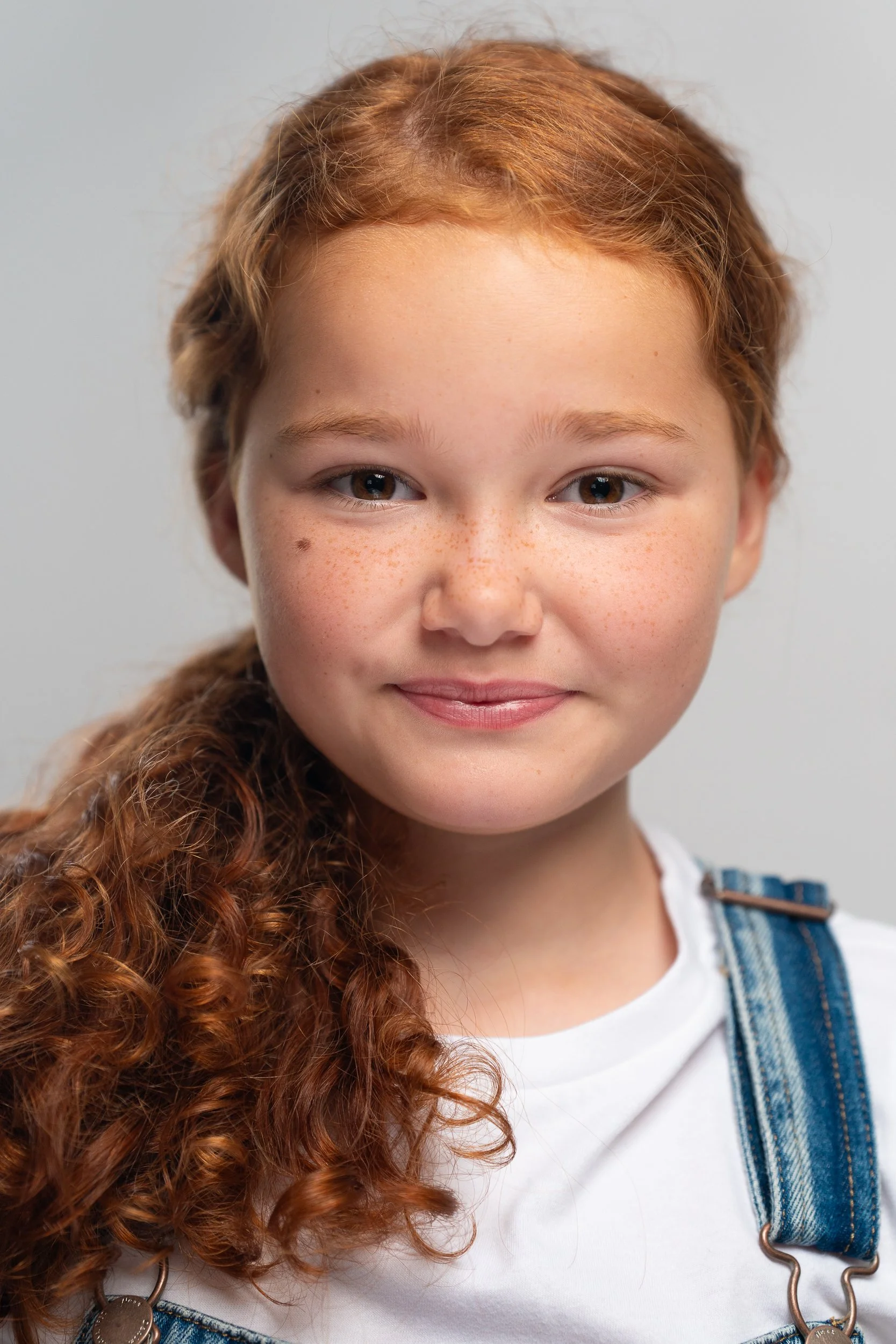 Close-up portrait of a young girl with long, curly red hair and freckles, wearing a white shirt and denim overalls, smiling softly against a plain background.