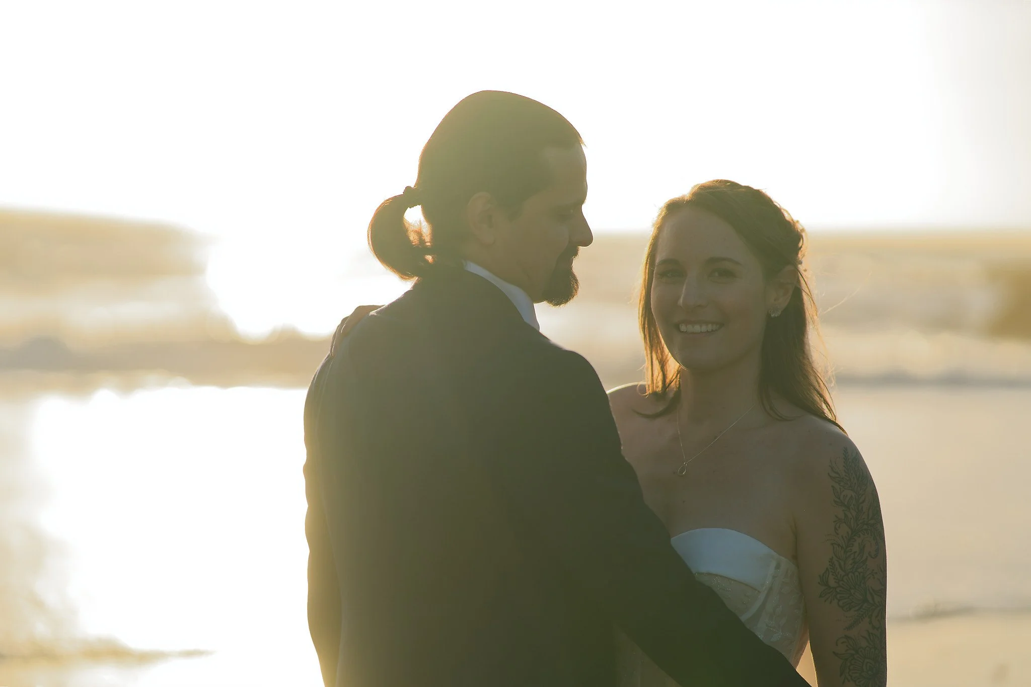 A couple dressed in wedding attire standing on the beach at sunset, with the man in a black suit and the woman in a strapless wedding gown, smiling at each other.