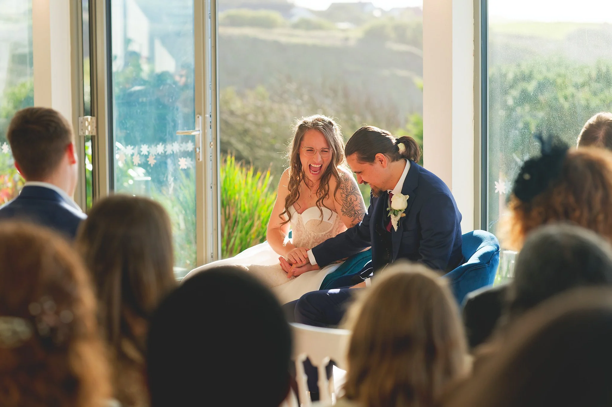 A bride and groom sharing a joyful moment during their wedding ceremony, sitting close with the bride laughing and holding the groom's hand, surrounded by guests.