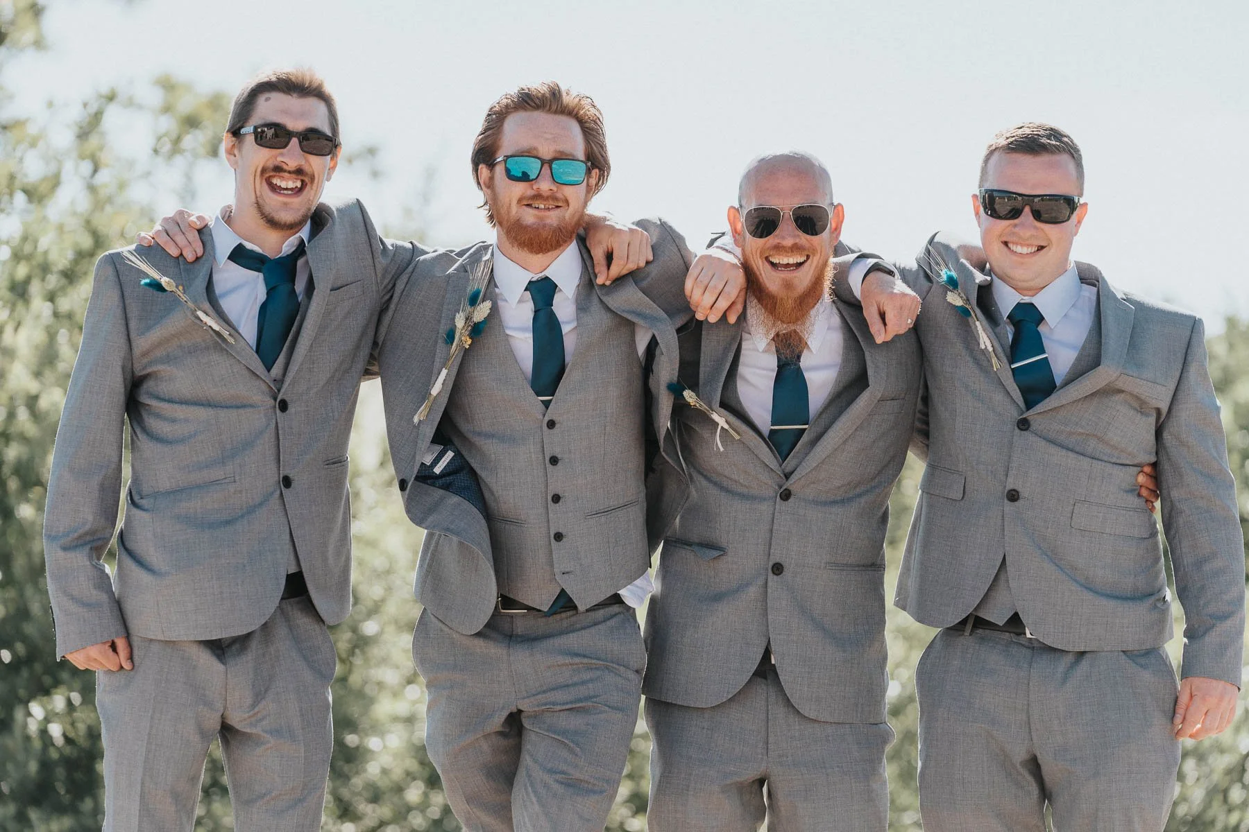 Four men in gray suits and sunglasses, arms around each other, smiling outdoors on a sunny day.