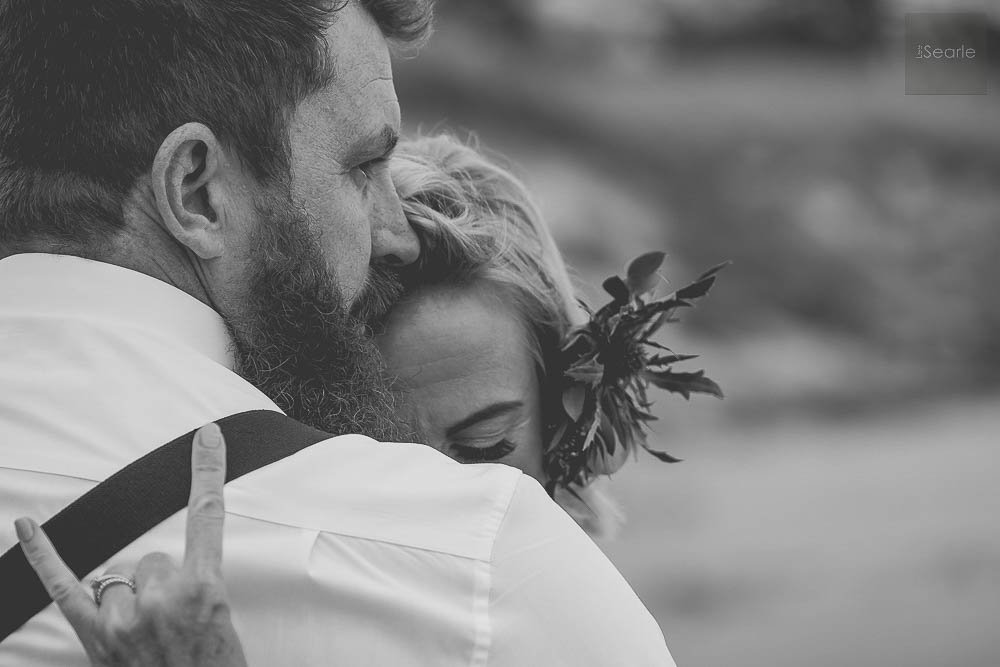 A man and woman sharing an embrace, with the man's face close to the woman's head, holding her close. The woman has a floral headpiece and a joyful expression. The photo is in black and white.