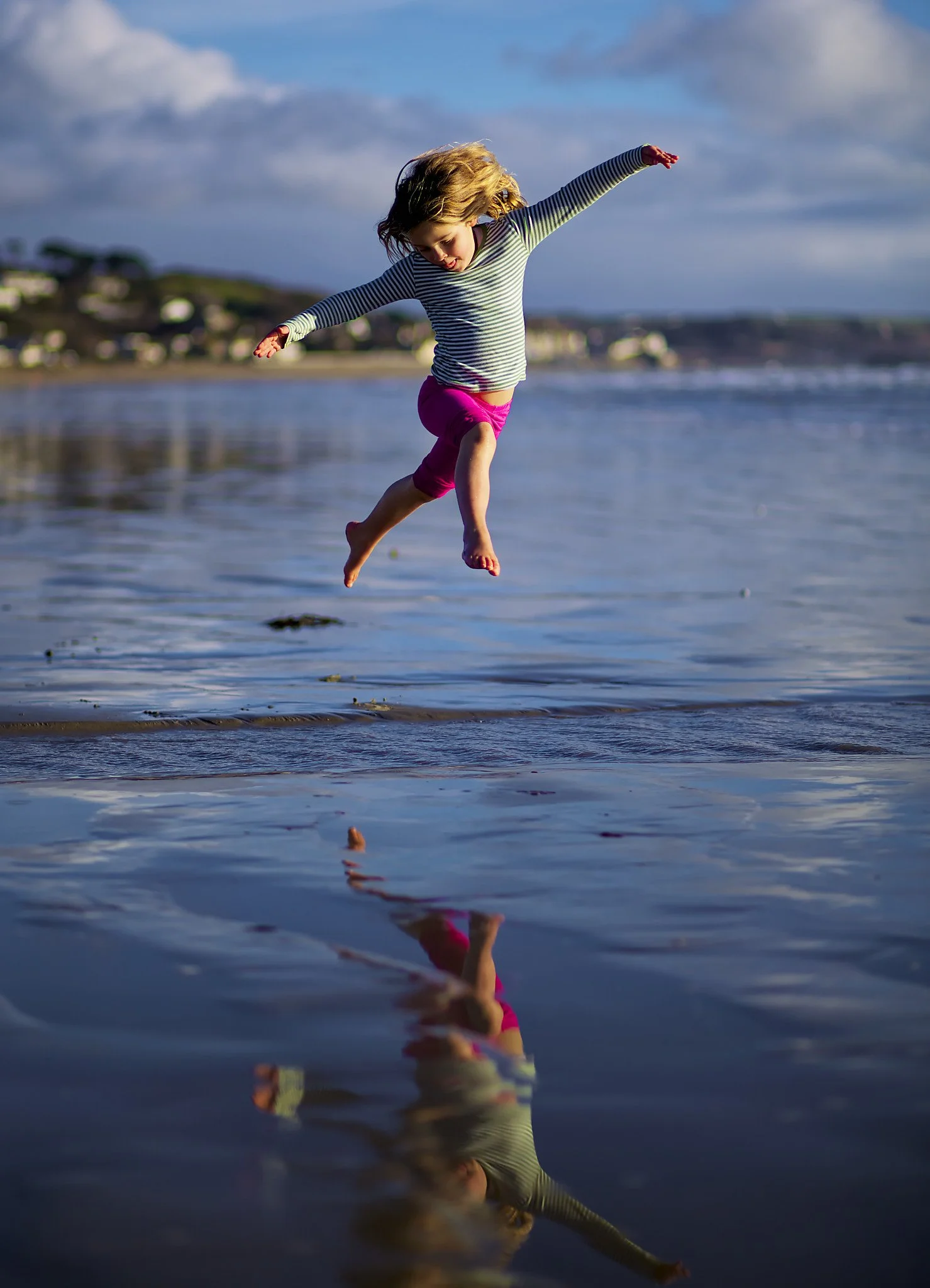A young girl with blonde hair wearing a striped long-sleeve shirt and pink shorts is jumping into the water at the beach, with her reflection visible on the surface.