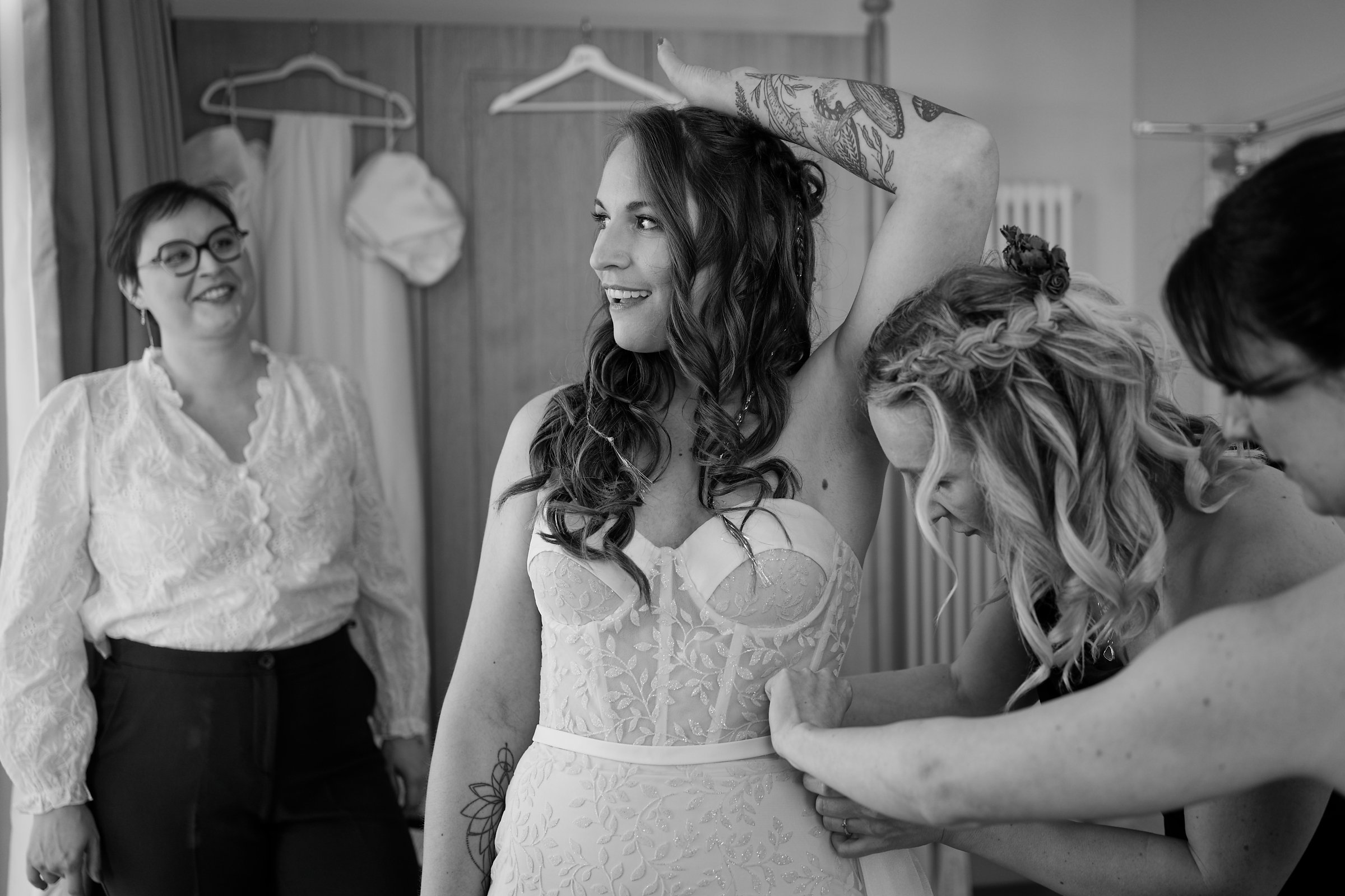 A bride in a lace wedding dress being helped by her bridesmaids in a dressing room.