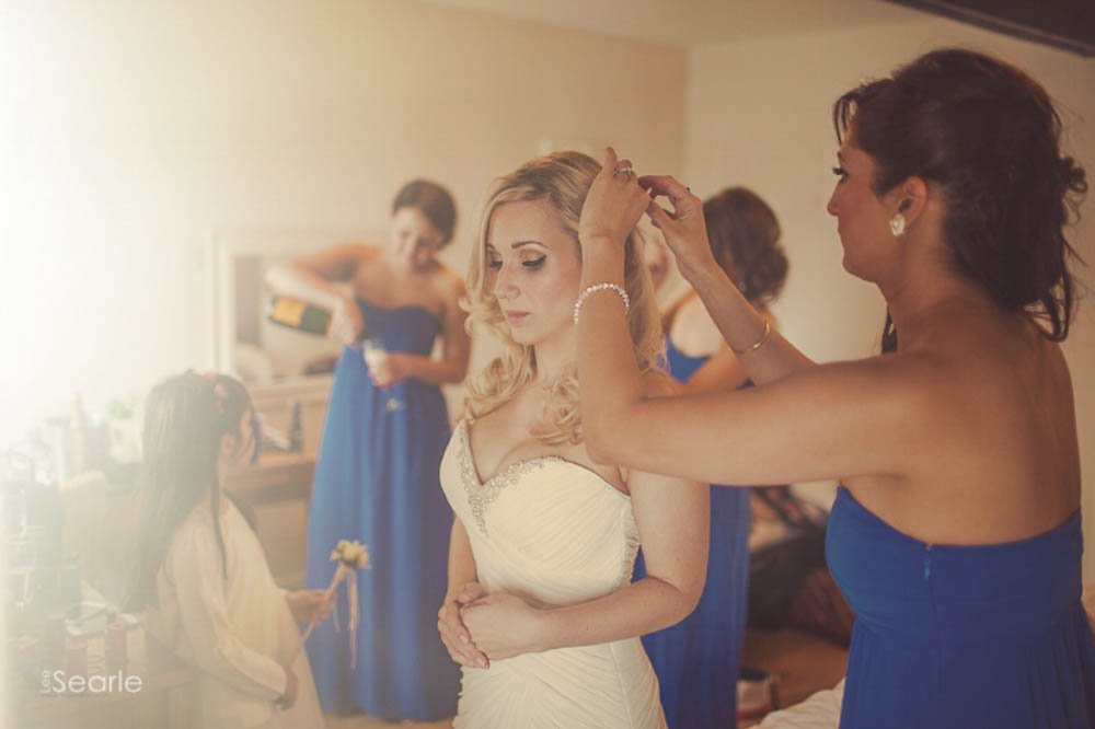 Bride getting her hair styled by a woman, with bridesmaids in blue dresses in the background.