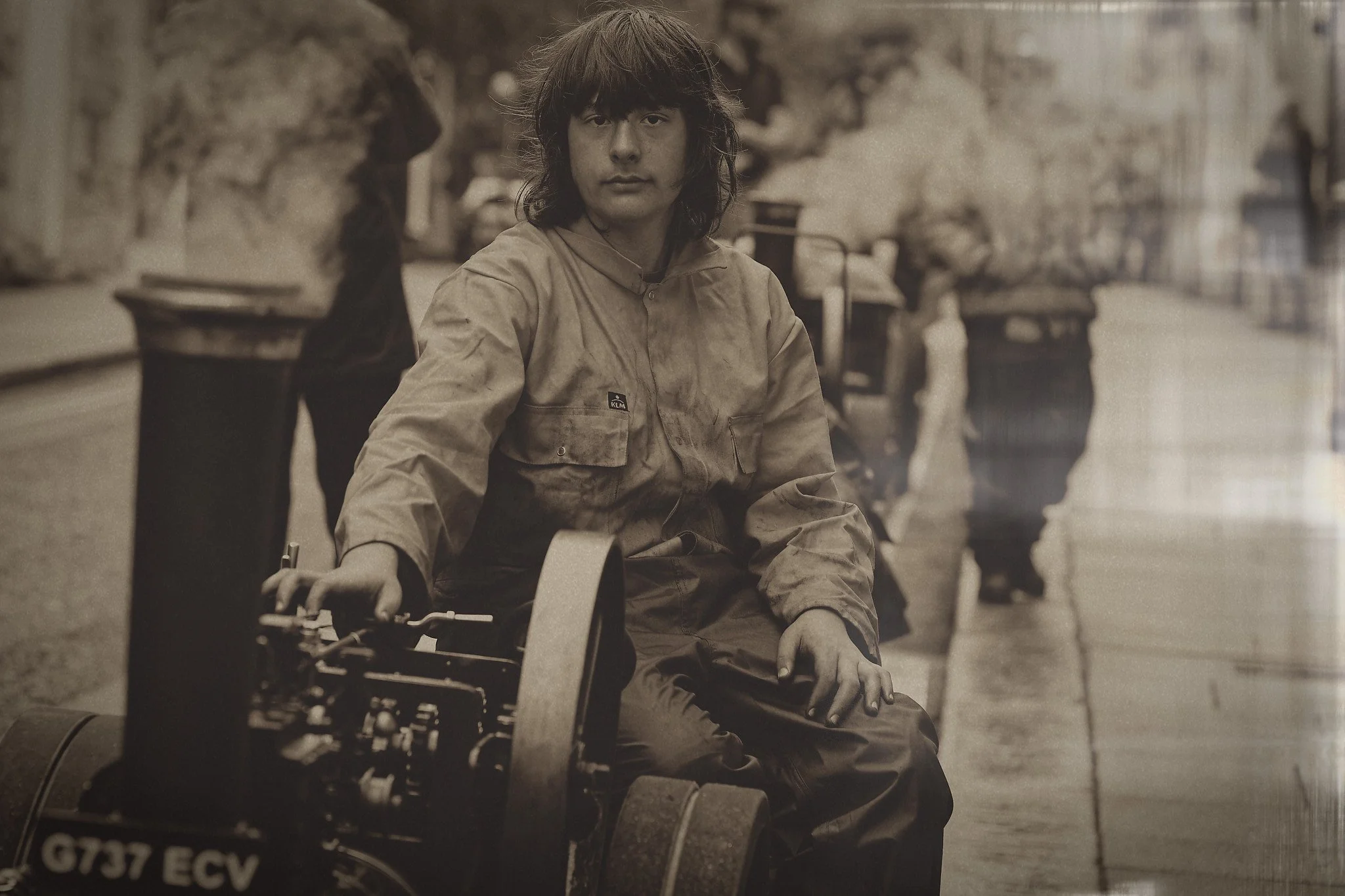 A young boy with long hair sitting on a miniature vehicle in the street, with a blurry background of a sidewalk and outdoor scene.