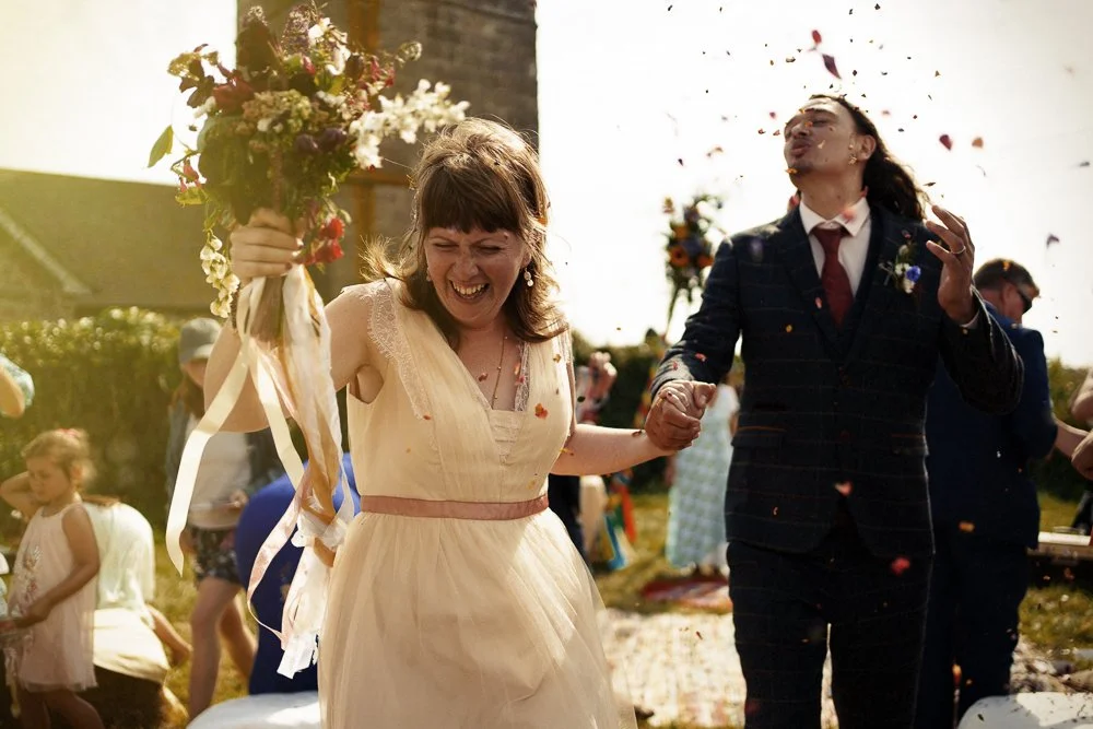 A woman in a light yellow dress holding a bouquet of flowers, smiling, and a man in a dark suit holding her hand, both celebrating outdoors amid fall confetti and other people.