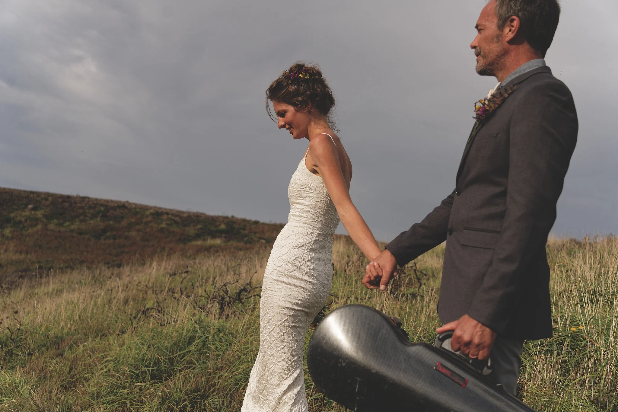 A bride in a white wedding dress and a groom in a grey suit holding hands, walking through a grassy field with a guitar case, under cloudy skies.