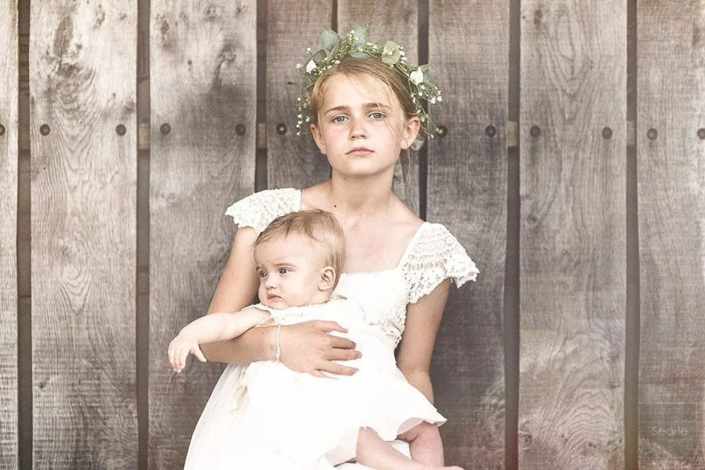 A young girl with a floral crown holding a baby girl, both dressed in white, standing in front of a wooden fence.
