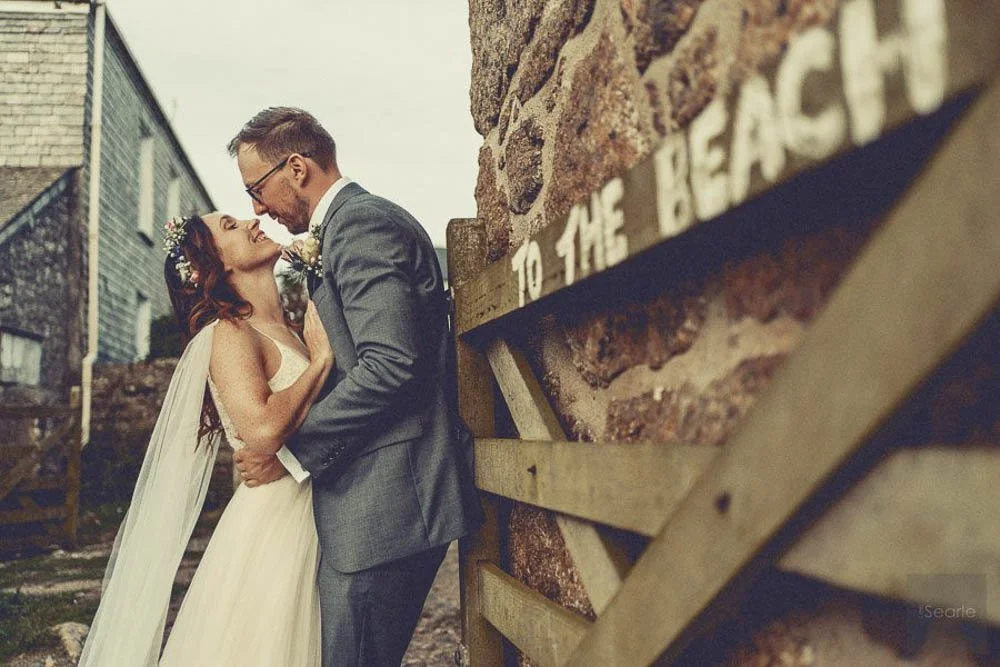 A bride and groom share an intimate moment outdoors near a brick wall, with the bride wearing a white wedding dress and flowers in her hair, and the groom wearing a gray suit with a boutonnière.