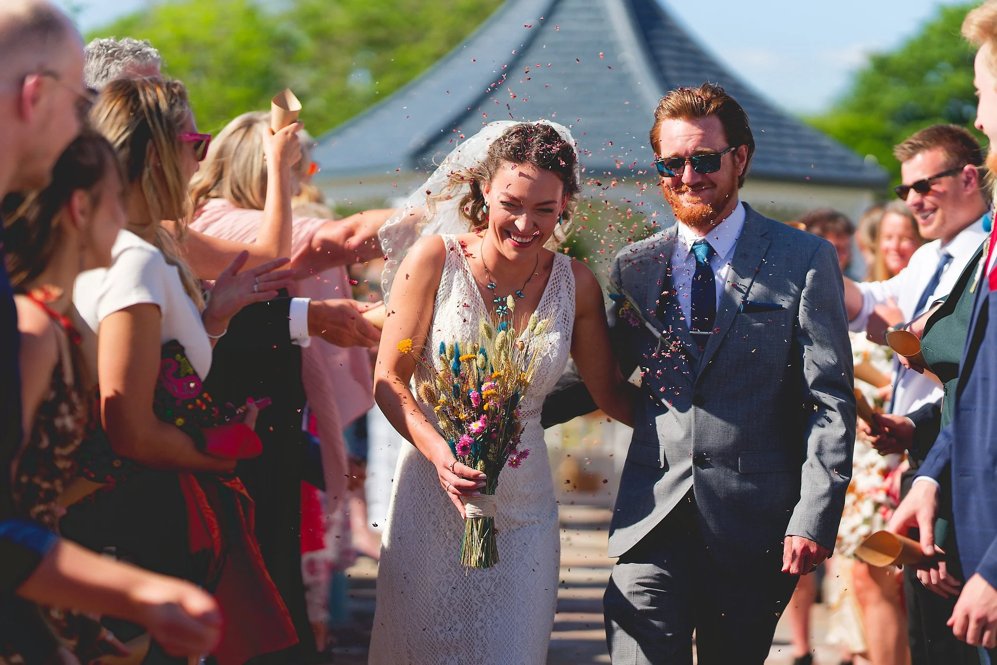 A bride and groom walk together smiling through a crowd throwing flower petals during their outdoor wedding celebration.