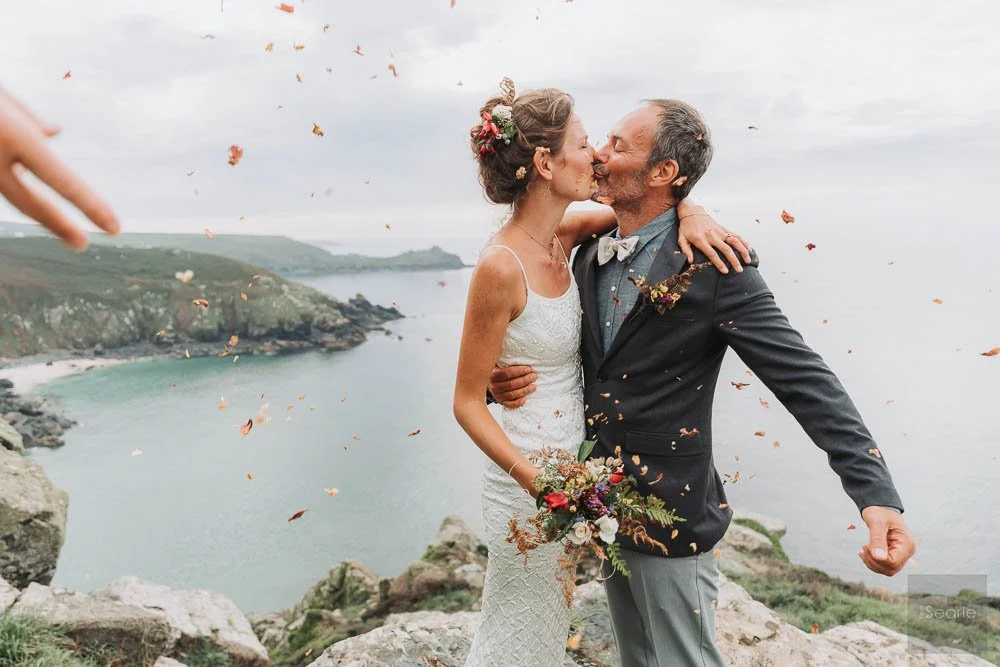 A couple in wedding attire kissing on a cliff overlooking the ocean with flowers and confetti.