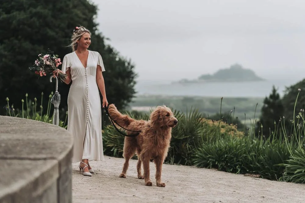 Woman in white dress holding a bouquet and leash, standing near a large brown dog on a leash, outdoors with greenery and a body of water in the background.
