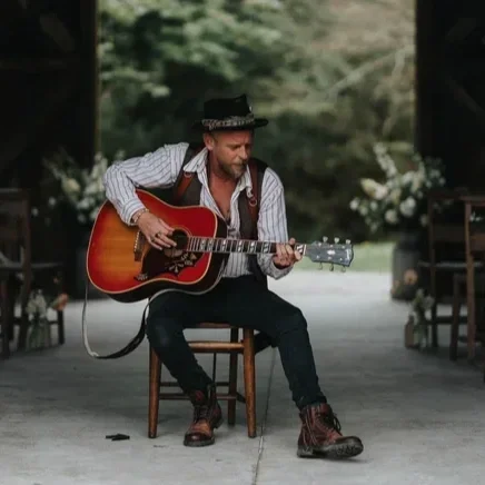 Lee Searle Photography playing an acoustic guitar outdoors surrounded by trees.
