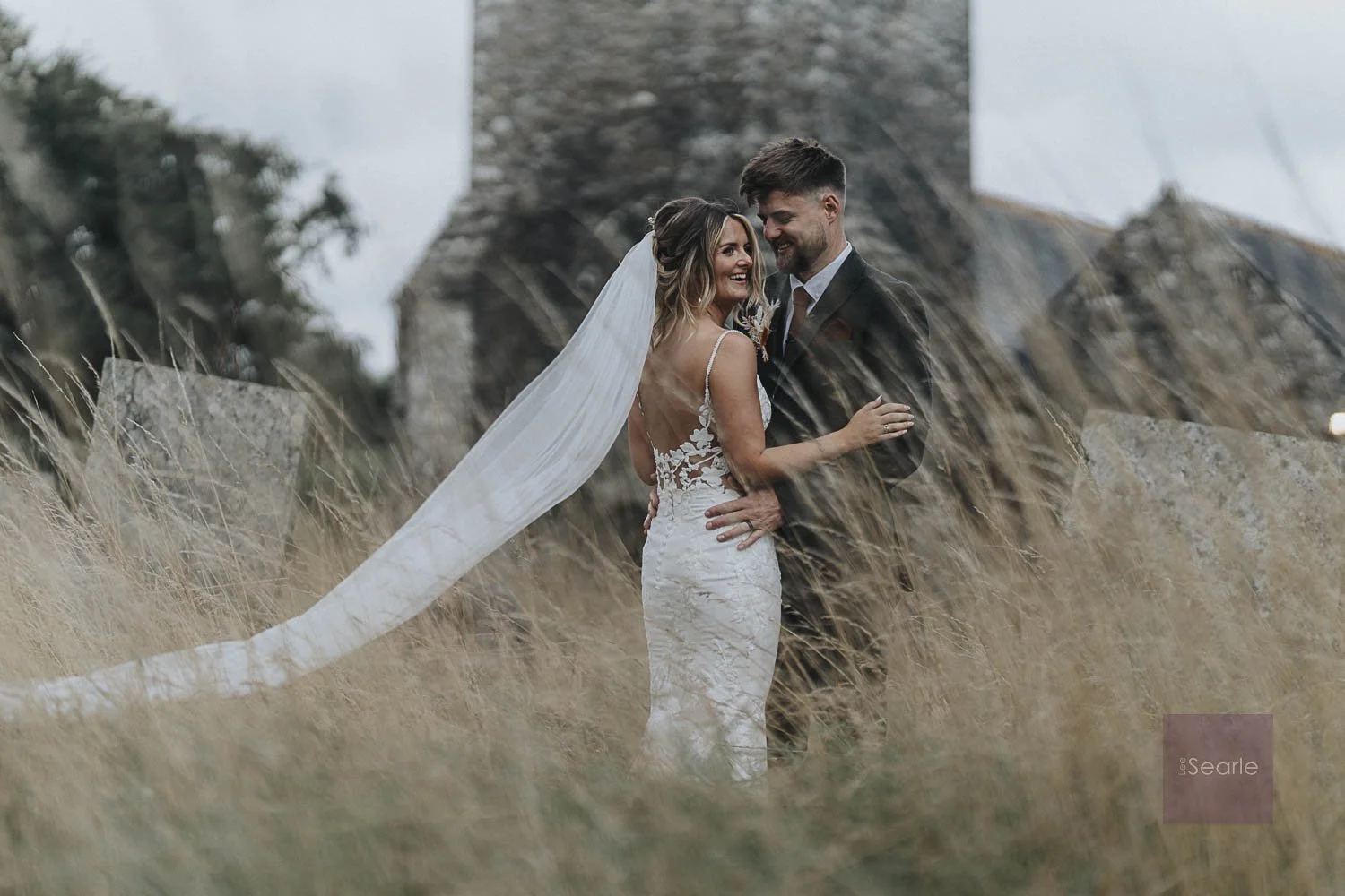 A bride and groom smiling and embracing outdoors, with dried grass in the foreground and a stone building in the background.