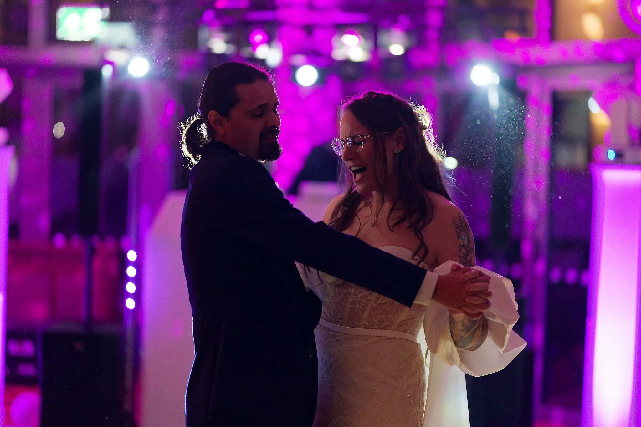 A couple dancing together at a wedding reception, with purple lighting in the background.