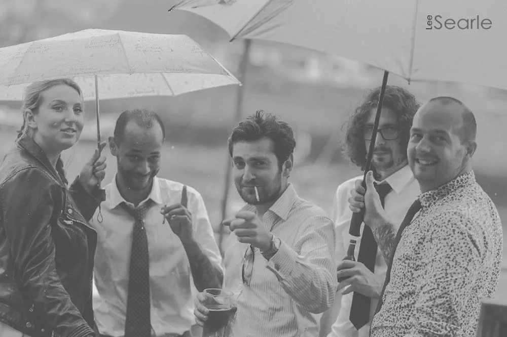 A group of five people, three men and two women, standing outdoors in the rain with umbrellas, some holding drinks, and one smoking a cigarette, in black and white.