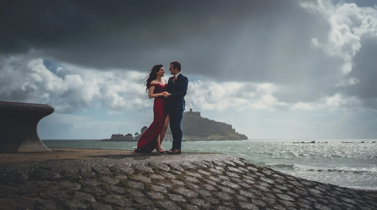 A couple dressed in formal attire standing on a cobblestone promenade near the ocean, with an island and castle in the background under cloudy skies.