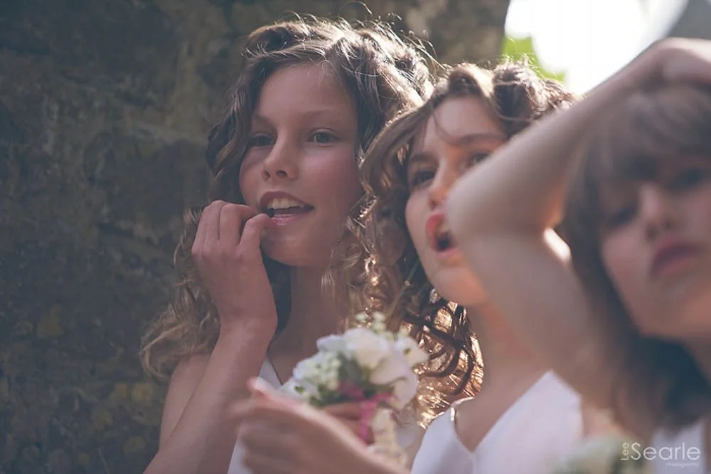 Three young women with curly hair, dressed in white, gathered outdoors with one holding a small bouquet of flowers, looking at something off-camera.