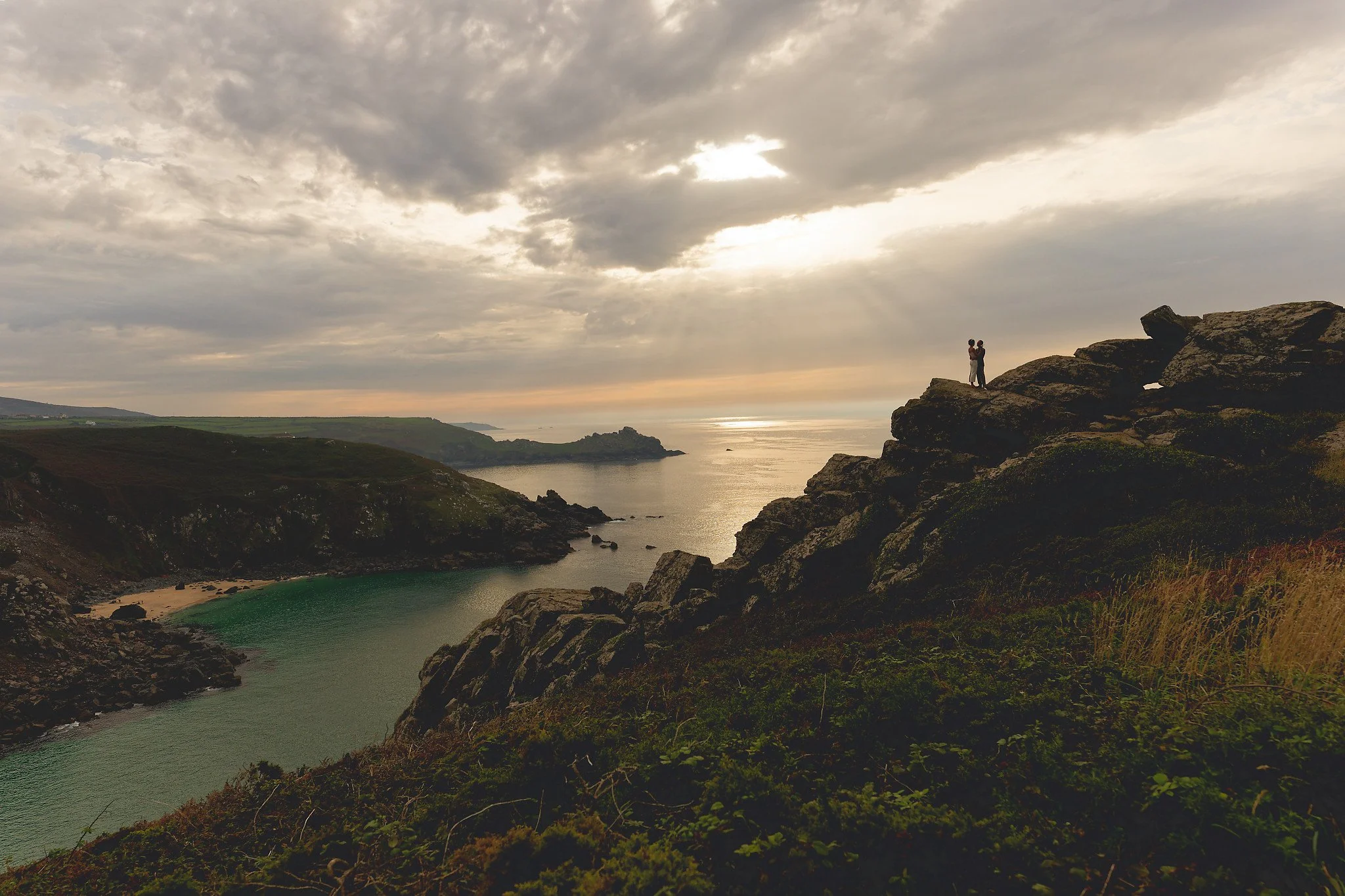 A couple standing on a rocky cliff overlooking a coastal cove with a sandy beach and green waters, under a cloudy sky with some sunlight breaking through.