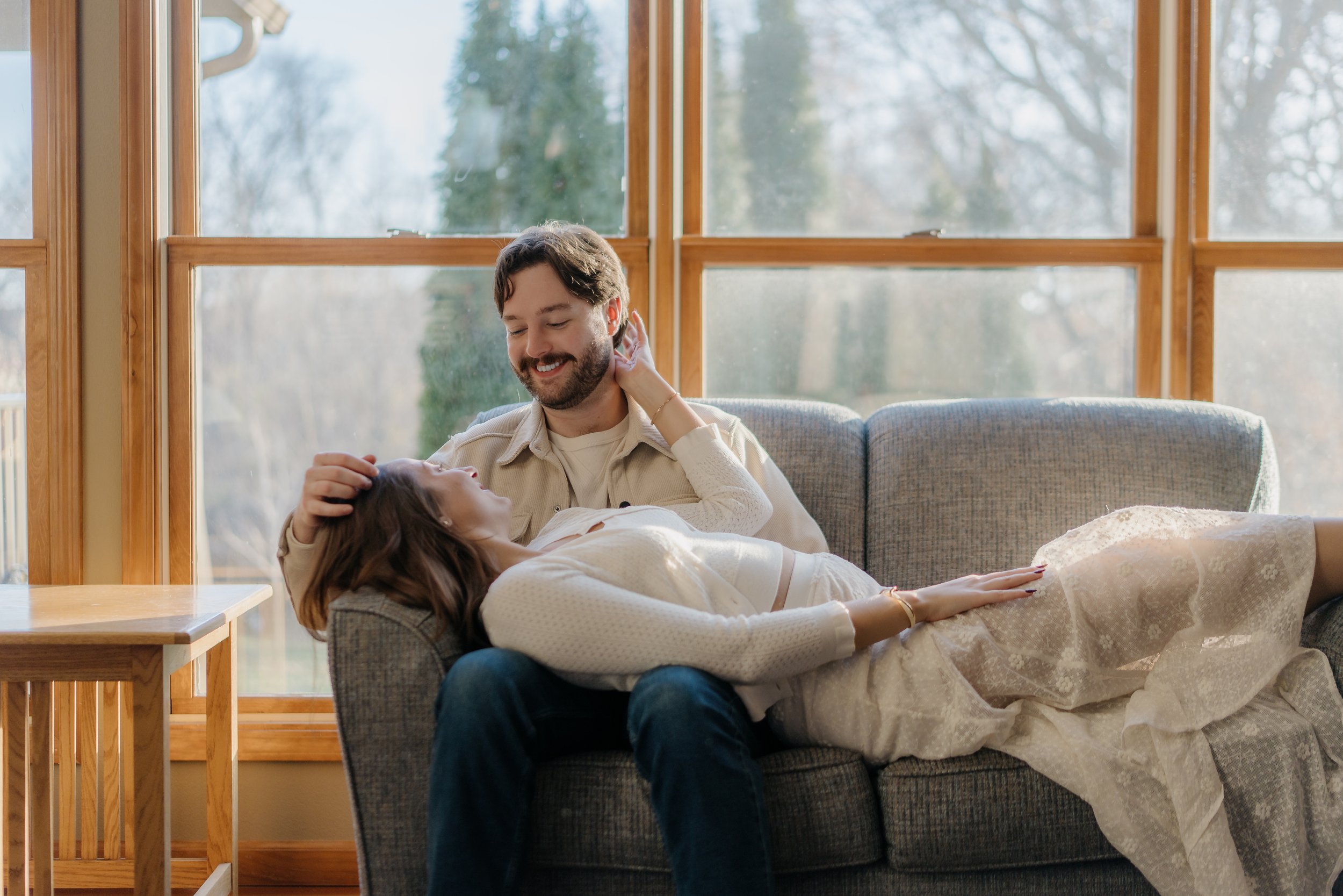 A man and a woman relaxing together on a sofa next to large windows with a view of trees outside, with the woman lying down and the man sitting beside her.